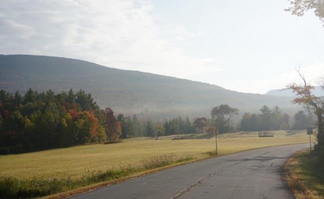 A road next to a meadow and tree-covered hill.