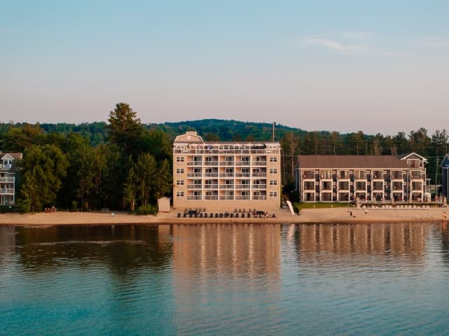 A waterfront view of the Alexandra Inn in Traverse City, Michigan, with its multi-story building and balconies overlooking a sandy beach and calm lake, surrounded by trees and hills in the background.