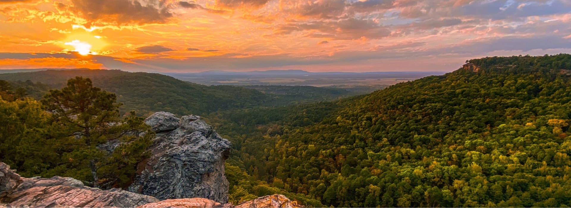 Sunset view over the Ozark Mountains with rolling forested hills, rocky overlooks, and warm light stretching across the landscape.