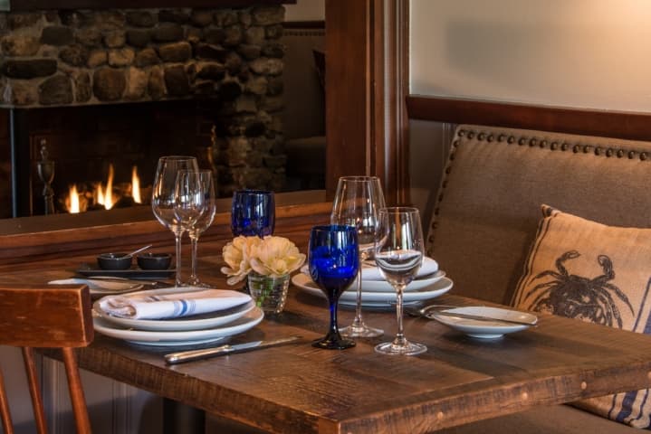 A view of a table set with blue water glasses, wine glasses, place settings and flowers.  A fireplace burns in the background envoking a cozy atmosphere.