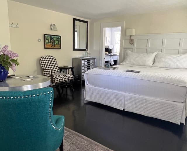Bedroom with white linens and white headboard, a black & white chair, mirror bureau and door leading to a bathroom