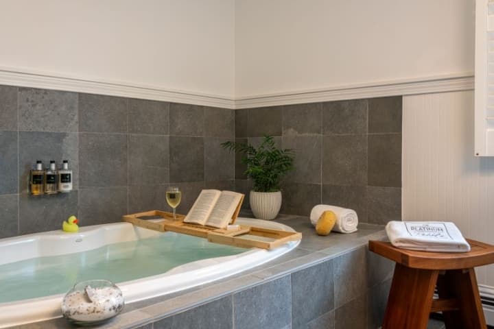 A bathtub with a book, glass of wine on a shelf above the tub, a plant and towels nearby