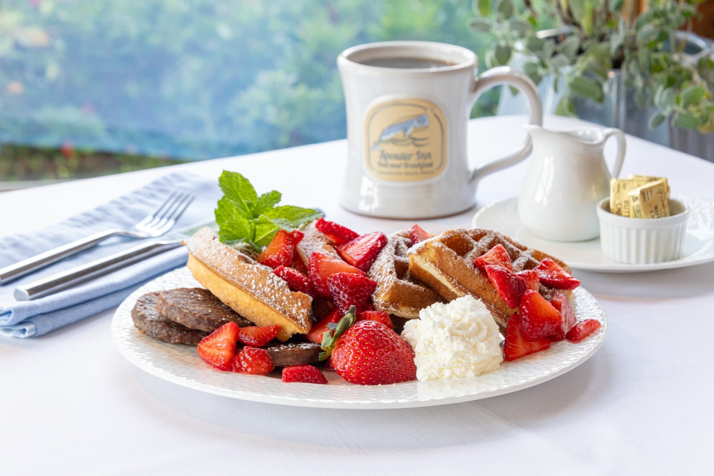 A plate of waffles, berries, and cream set on a table with white tablecloth, along with a set of silverware, mug of coffee, creamer, and butter.