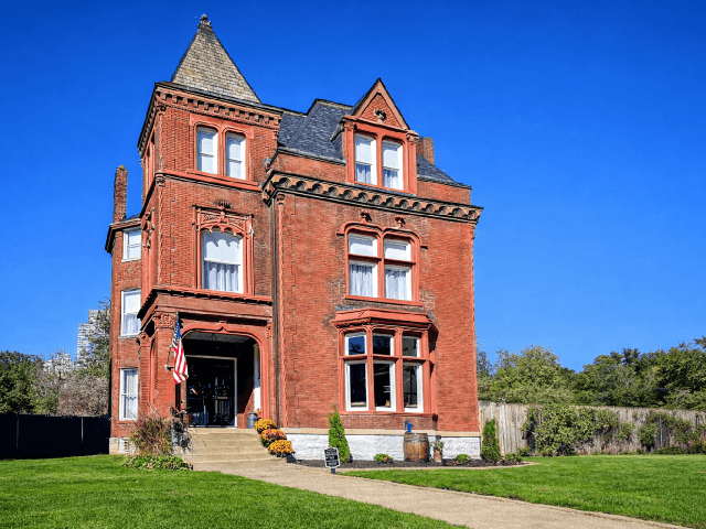 Red brick Victorian mansion with ornate trim and a turreted roofline, set on a manicured green lawn under a clear blue sky. An American flag hangs beside the covered front entrance, with a paved walkway leading to the front steps and bourbon barrels and s