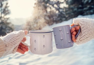 Man and woman hands in knitting mittens with cups of hot tea on winter forest glade
